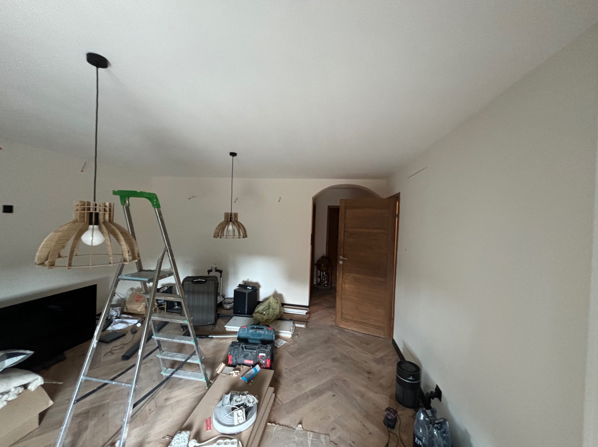 Wnętrze w trakcie remontu: drabina, narzędzia, osłonięte lampy, ściany przygotowane do malowania, parkiet w jodełkę, widoczne ślady po pracach.