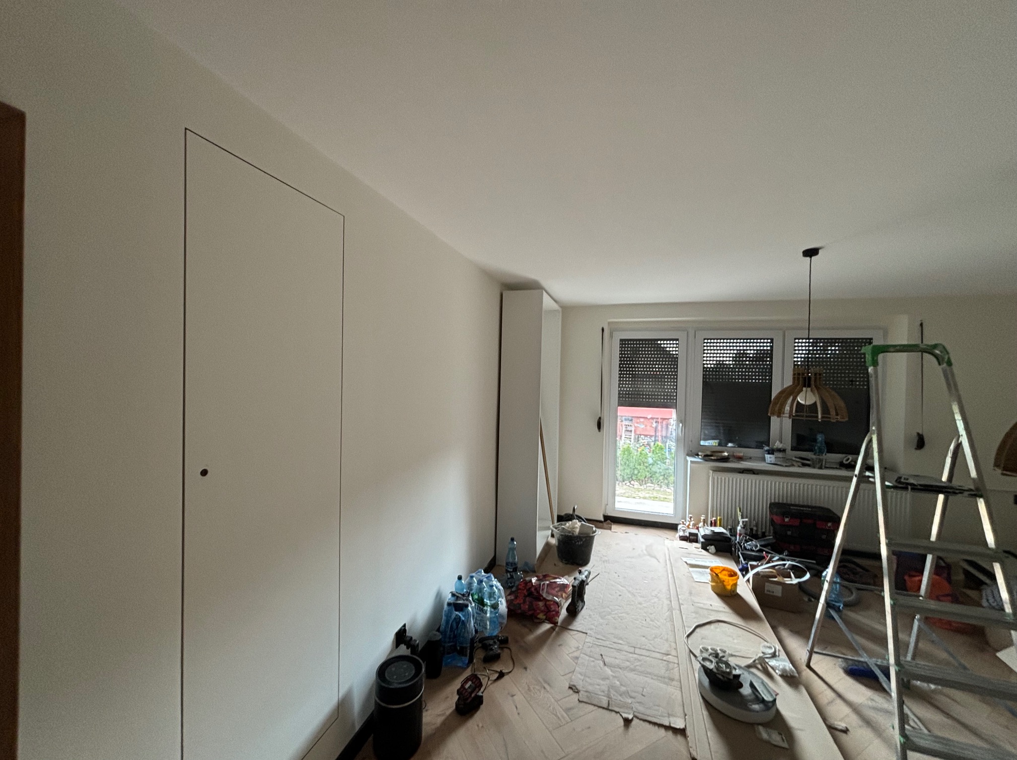 Pokój w trakcie remontu: pomalowane ściany na biało, zabezpieczona podłoga, drabina, narzędzia i materiały malarskie widoczne w pomieszczeniu.