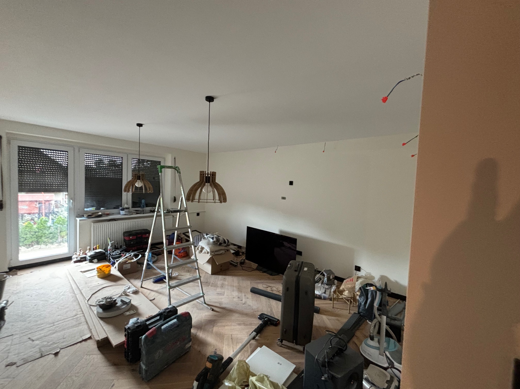 Pokój w trakcie remontu: drabina, narzędzia, lampy wiszące i świeżo pomalowane ściany z widocznymi otworami na kable. Podłoga parkietowa częściowo zakryta materiałami zabezpieczającymi.