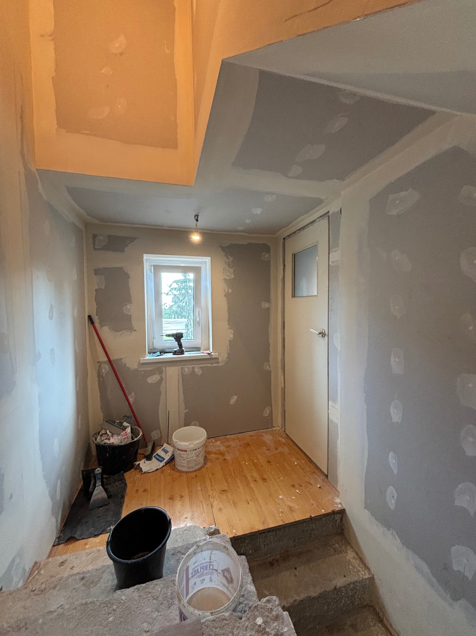 Wnętrze w trakcie remontu: ściany częściowo pokryte szpachlą, widoczne narzędzia i wiaderka z zaprawą, drewniana podłoga zabezpieczona przed zabrudzeniem, okno z widokiem na zieleń.