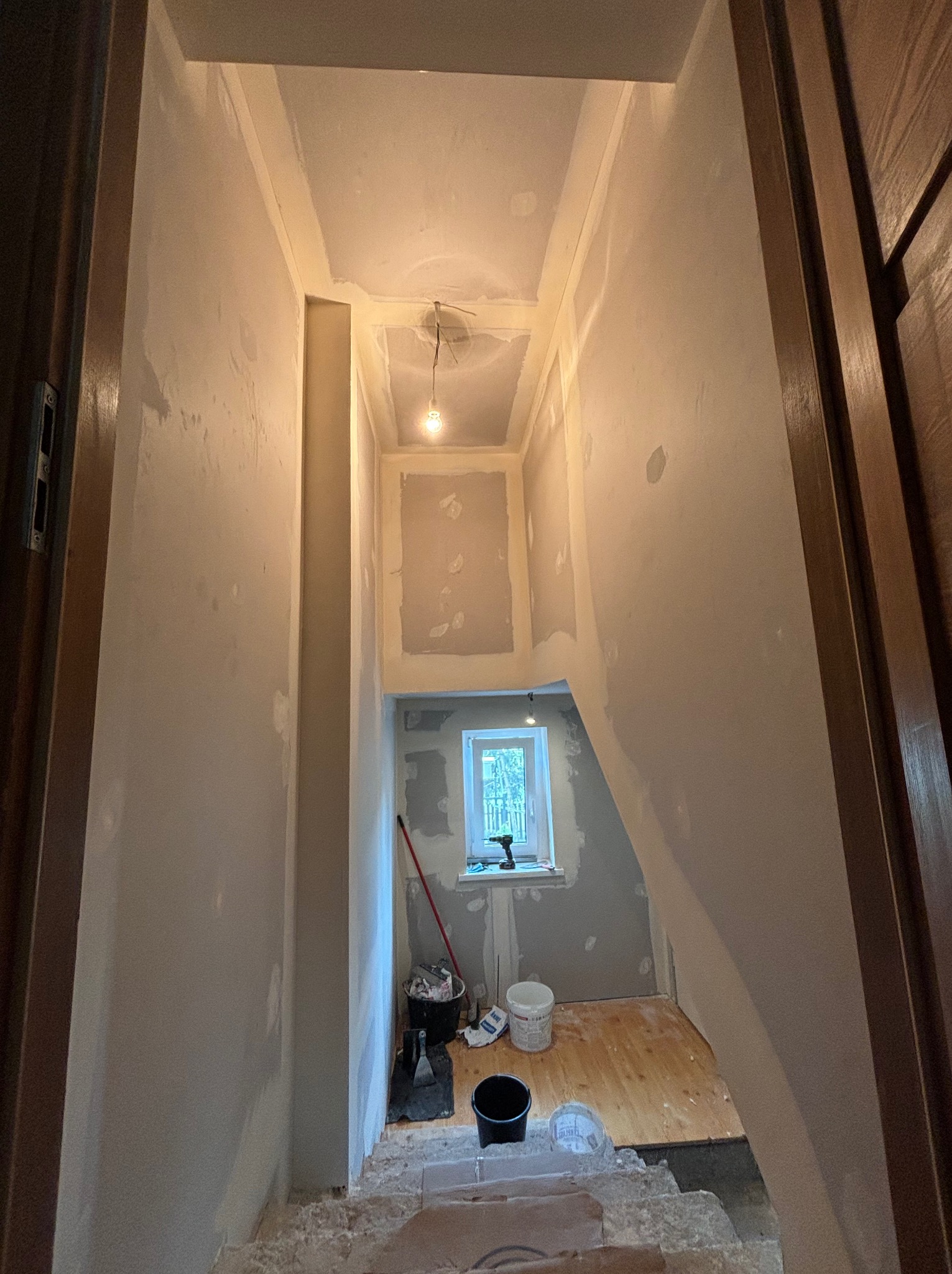 Korytarz w trakcie remontu, ściany częściowo pokryte masą szpachlową, widoczne narzędzia i wiaderka, surowe drewniane podłogi w dolnej części korytarza, okno w tle.