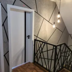 Nowoczesne wnętrze: białe drzwi z czarnym akcentem, geometryczna tapeta, drewniana podłoga i czarna balustrada schodowa z designerskimi lampami wiszącymi.