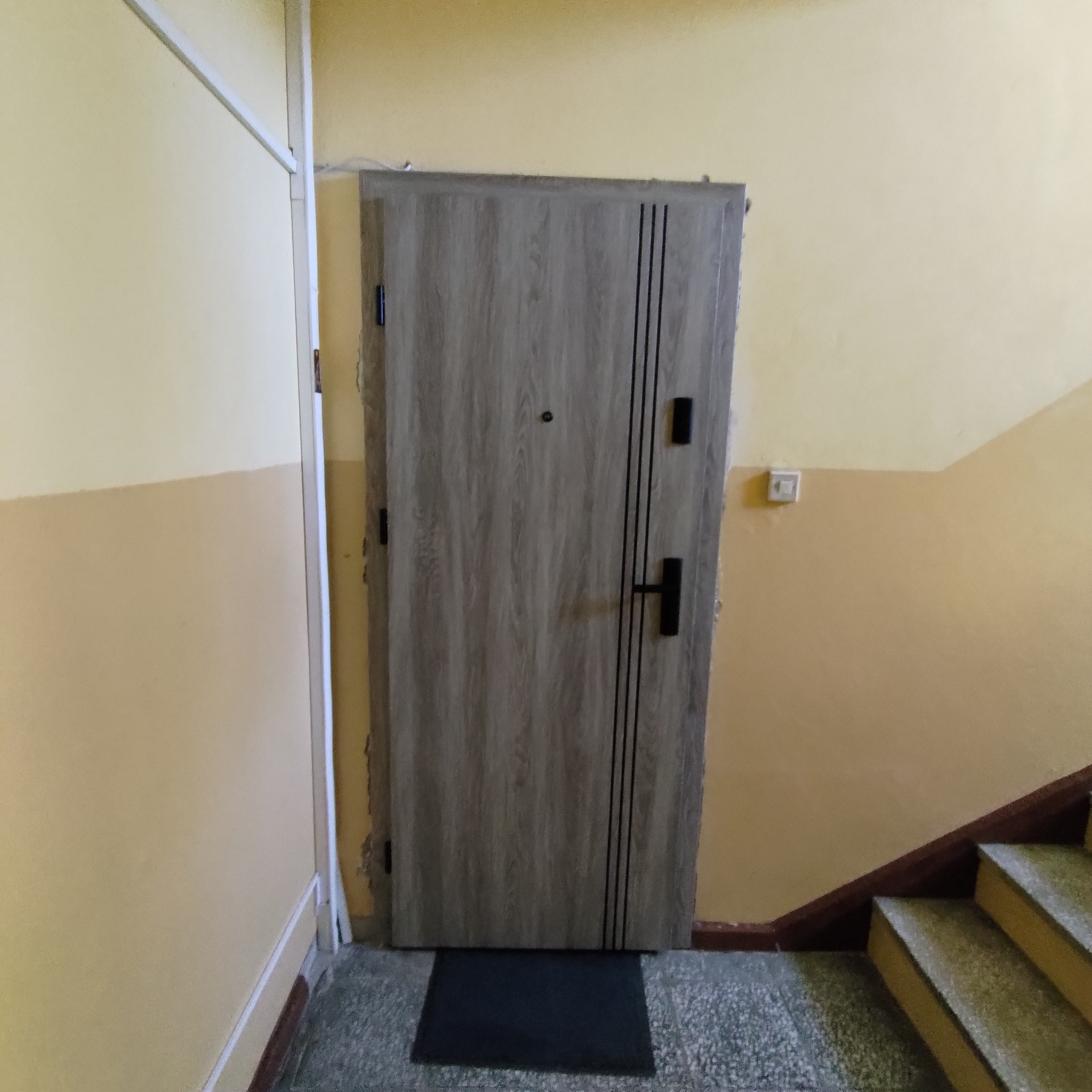 Szare drzwi wejściowe z czarnymi akcentami w klatce schodowej z żółtymi ścianami i szarymi schodami.