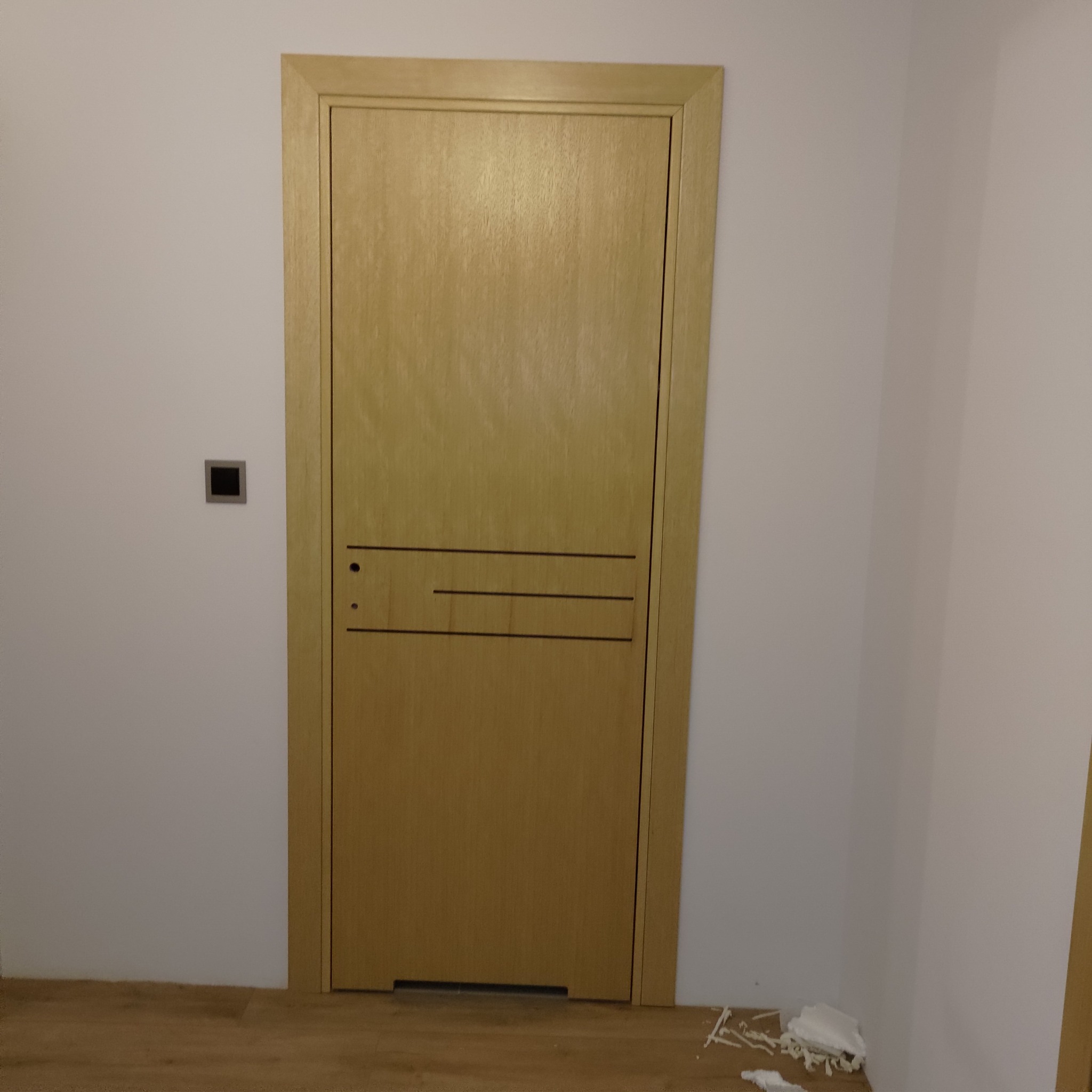 Zamknięte, drewniane drzwi wewnętrzne z prostokątnym panelem dekoracyjnym i dwiema poziomymi liniami, zamontowane w białej ścianie nad drewnianą podłogą, z widocznymi fragmentami styropianu...