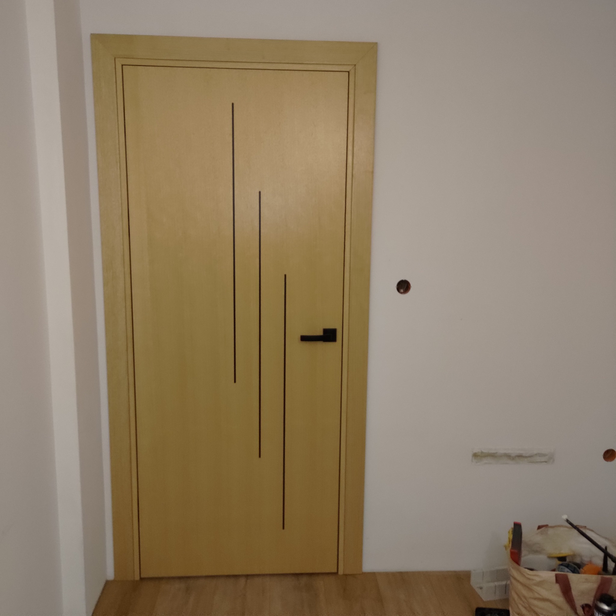 Zamontowane, jasne, drewniane drzwi wewnętrzne z czarną klamką i trzema pionowymi, ciemnymi paskami dekoracyjnymi, widoczne na tle białej ściany z niedokończonymi pracami wykończeniowymi...