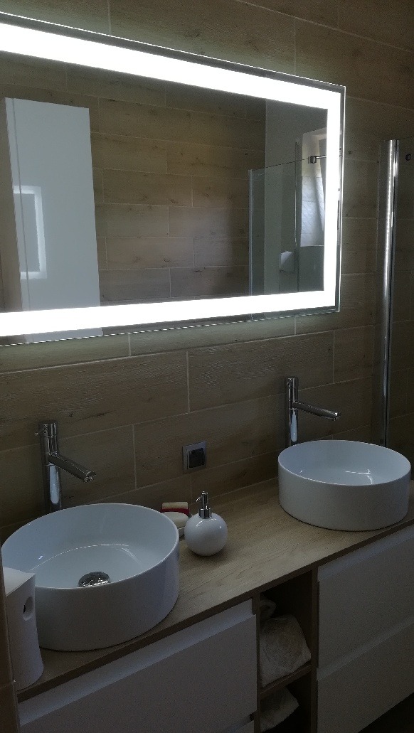 Nowoczesna łazienka z dwiema okrągłymi umywalkami nablatowymi, podświetlanym lustrem LED i płytkami imitującymi drewno na ścianie.