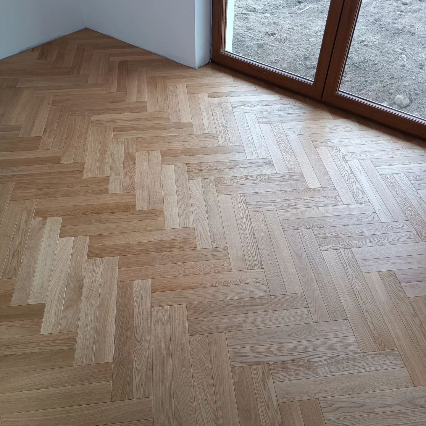 Jasna, drewniana podłoga ułożona w jodełkę, widok z góry, przy oknie i białej ścianie.