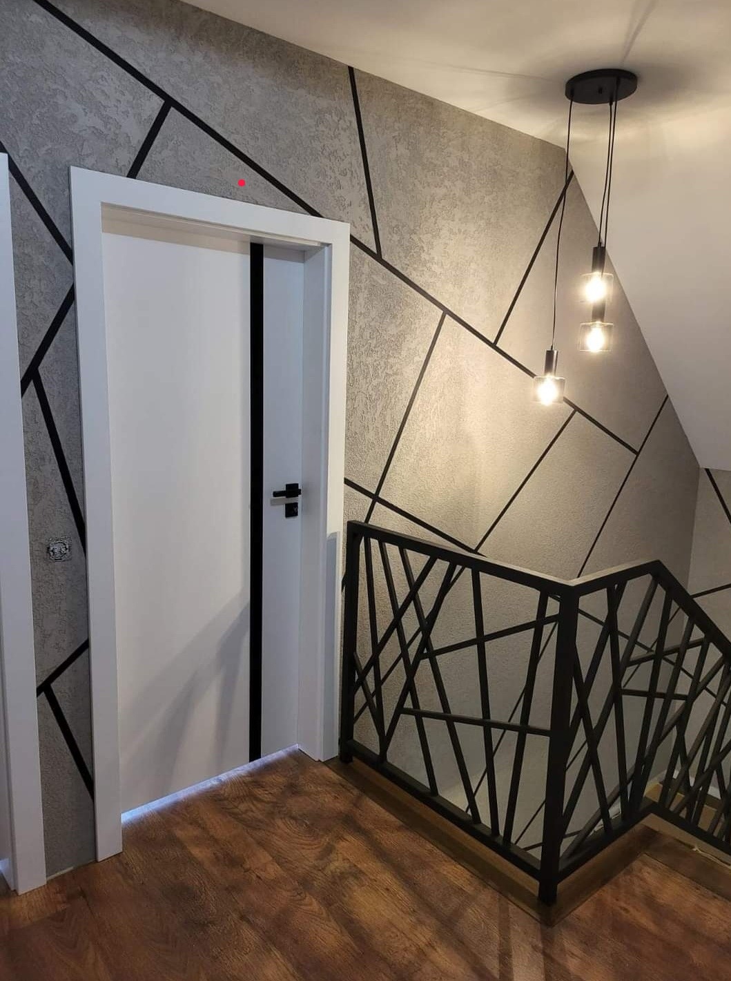 Nowoczesne wnętrze: białe drzwi z czarnym akcentem, geometryczna tapeta, drewniana podłoga i czarna balustrada schodowa z designerskimi lampami wiszącymi.