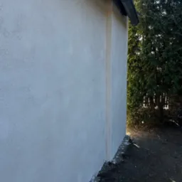 Ściana budynku po nałożeniu strukturalnego tynku zewnętrznego, widoczny fragment rynny i roślinność w tle.