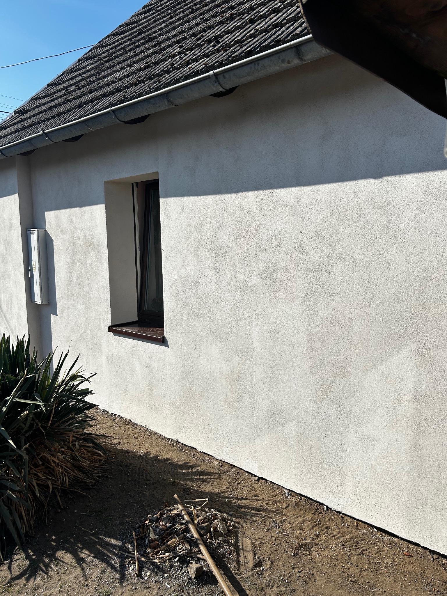 Świeżo otynkowana ściana budynku z widocznym oknem, skrzynką elektryczną i roślinnością u podstawy, z lekkim cieniowaniem i stertą gałęzi na ziemi.