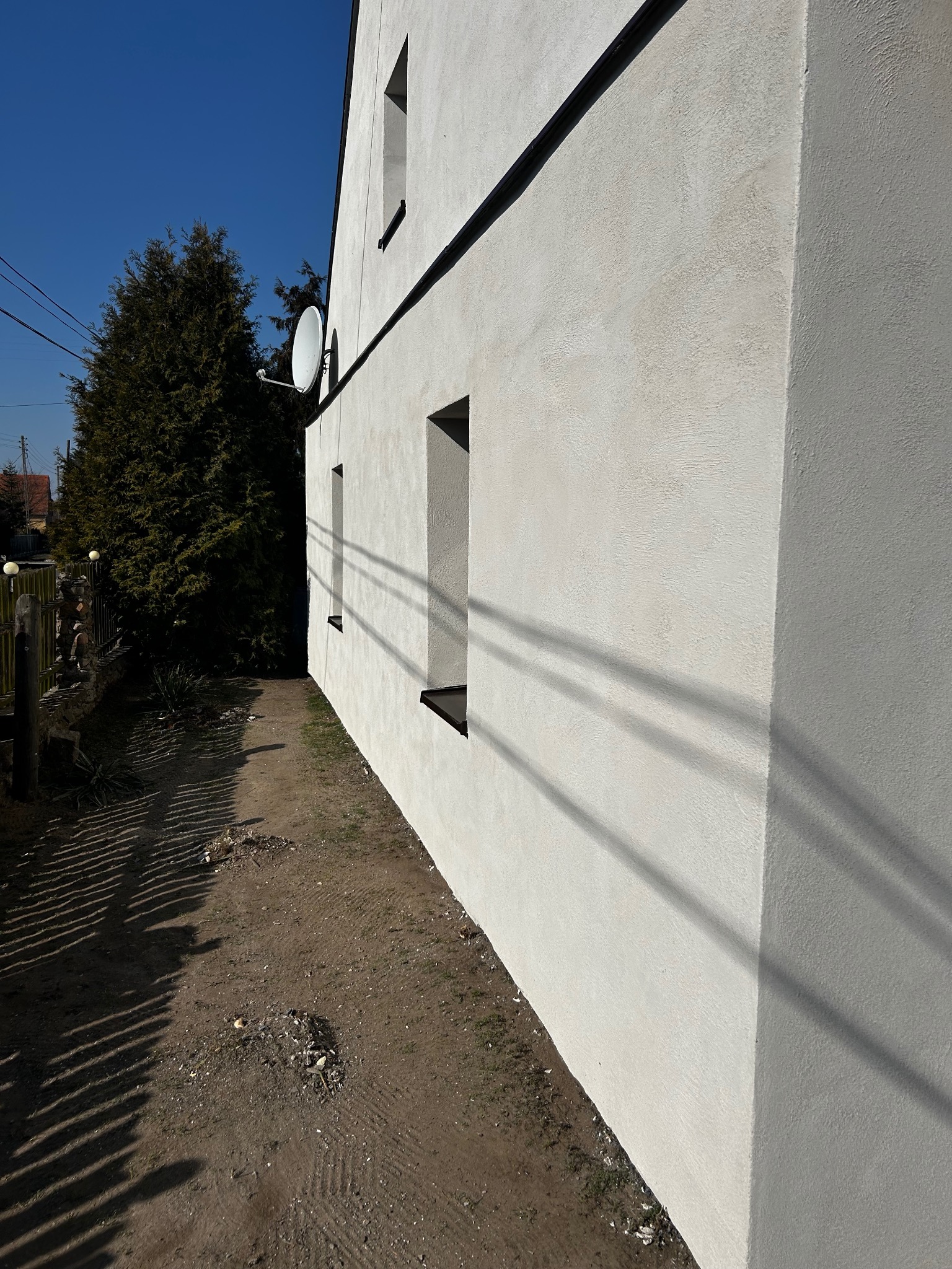 Świeżo otynkowana, jasna ściana zewnętrzna domu jednorodzinnego z widocznymi oknami i anteną satelitarną, z fakturą baranka w słoneczny dzień.