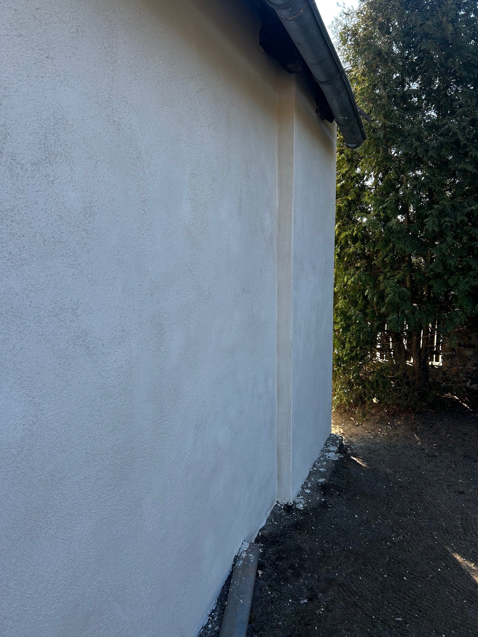Ściana budynku po nałożeniu strukturalnego tynku zewnętrznego, widoczny fragment rynny i roślinność w tle.