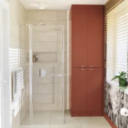 Jasna łazienka z kabiną prysznicową, szafką w kolorze terakoty i oknem z żaluzjami. Minimalistyczny design, płytki w odcieniach beżu, roślina w doniczce na parapecie.