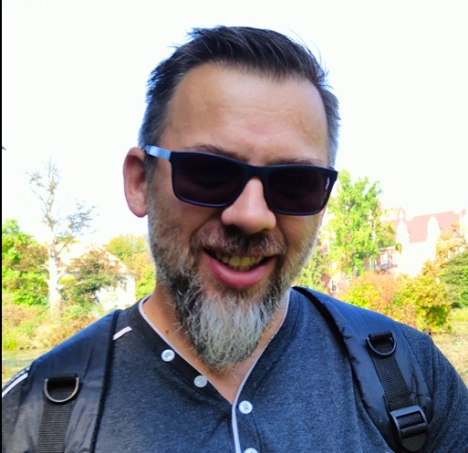 Portret uśmiechniętego mężczyzny w okularach przeciwsłonecznych z szpakowatą brodą, w tle zieleń i budynek z czerwonym dachem.