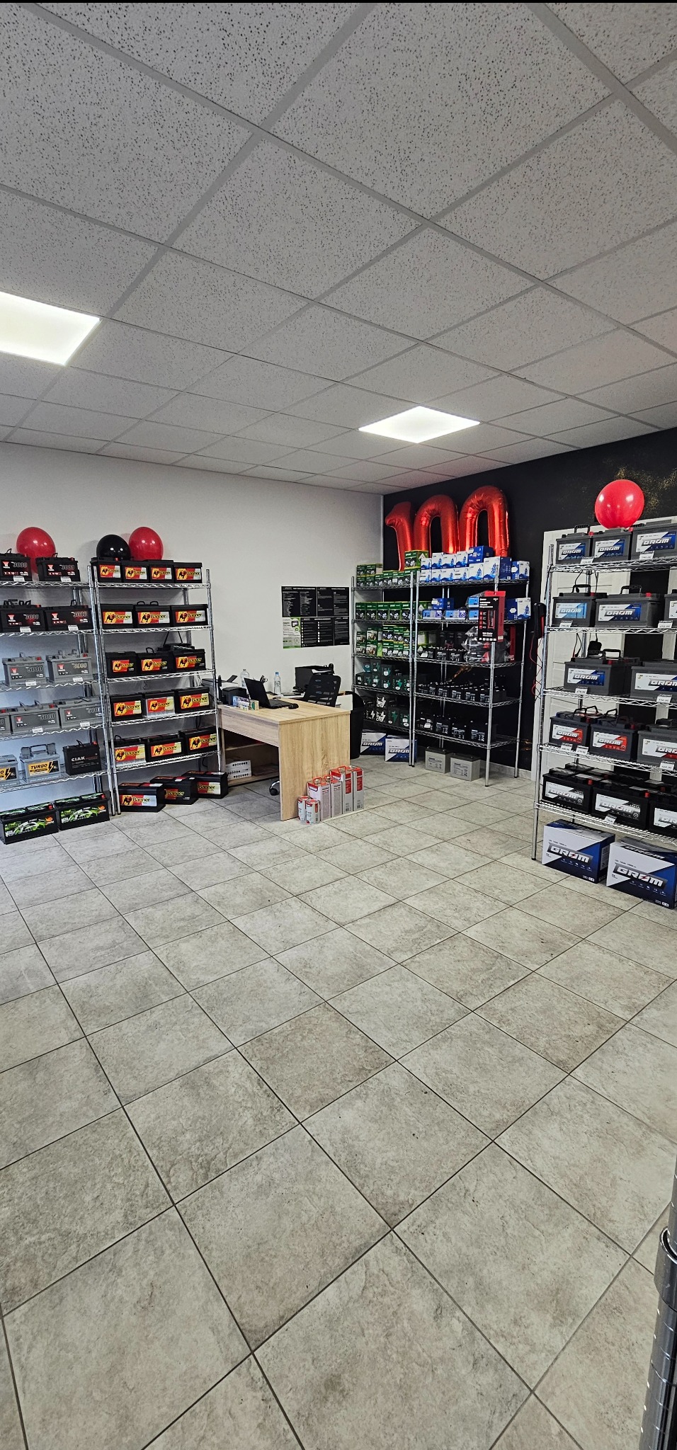 Wnętrze sklepu z akumulatorami samochodowymi, regały wypełnione produktami, czerwone balony i cyfra '100' z balonów, jasne płytki na podłodze, biurko w tle.