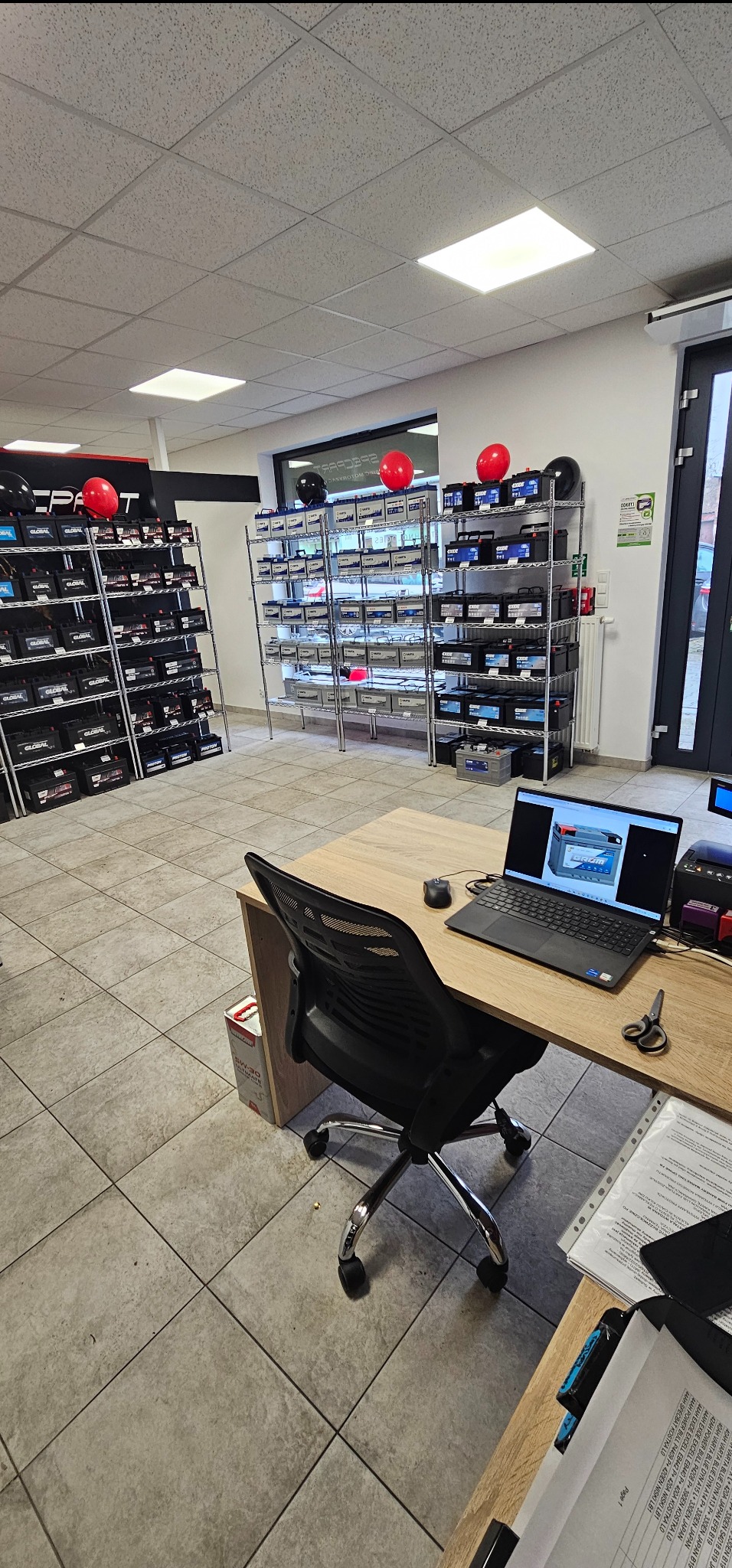 Wnętrze sklepu z akumulatorami samochodowymi: Rzędy półek z akumulatorami różnych marek, w tle biurko z laptopem wyświetlającym zdjęcie akumulatora, czerwone balony dekoracyjne.