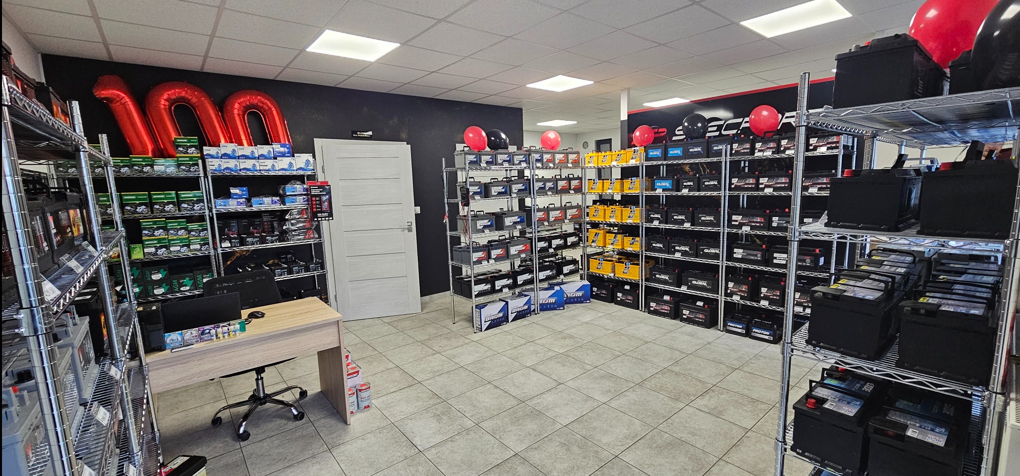 Wnętrze sklepu z akumulatorami samochodowymi, regały wypełnione różnymi modelami, dekoracje balonowe w tle, biurko z komputerem po lewej stronie.