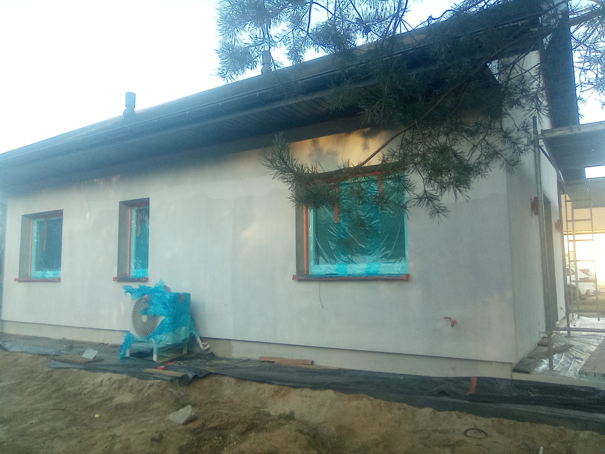Świeżo otynkowana elewacja domu jednorodzinnego z zabezpieczonymi oknami folią, wentylator budowlany owinięty folią ochronną, widoczne przygotowania do dalszych prac.