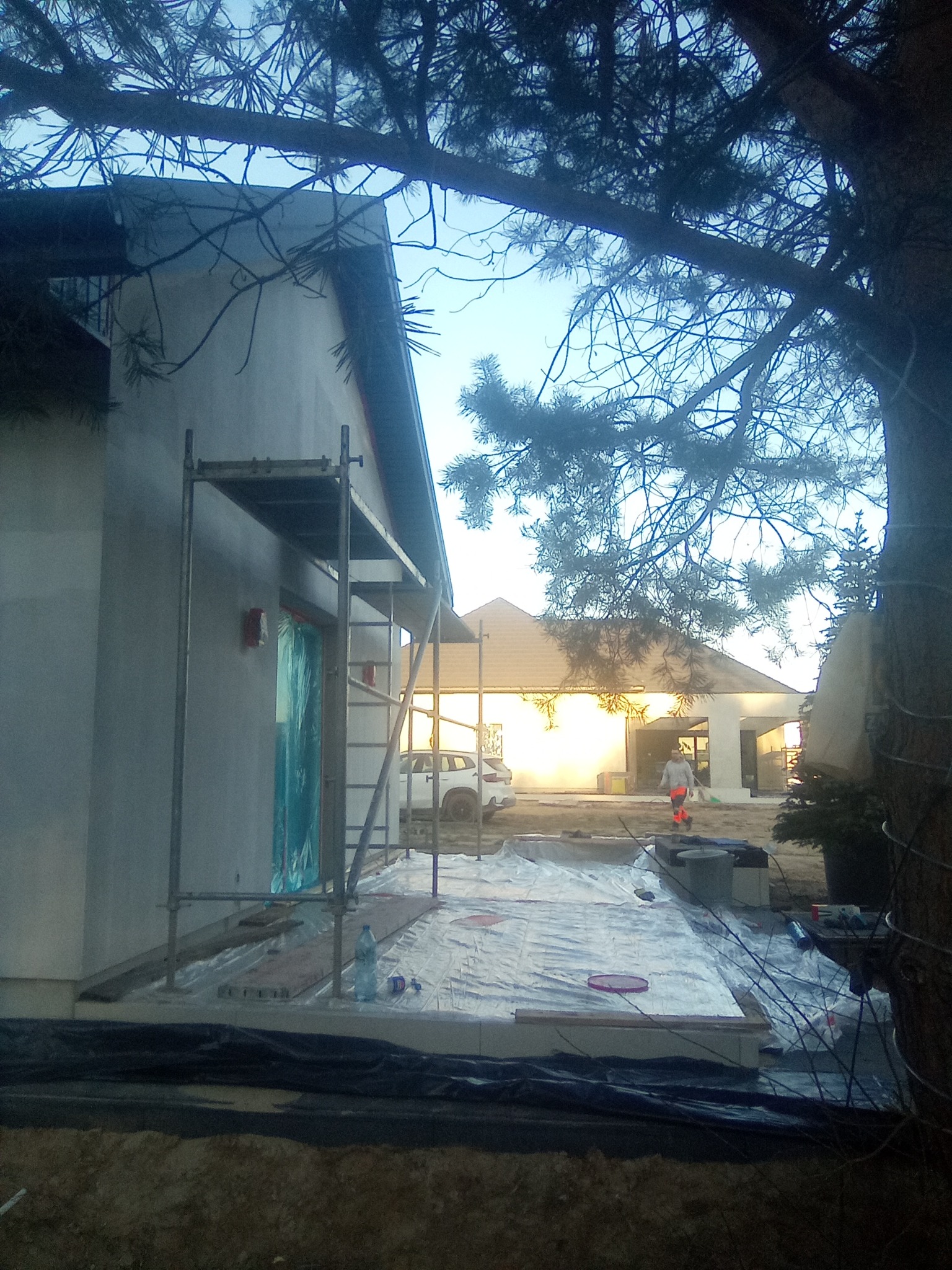 Dom w trakcie prac elewacyjnych, widoczne rusztowanie, zabezpieczona folią podstawa, świeżo otynkowana ściana i pracownik w oddali.