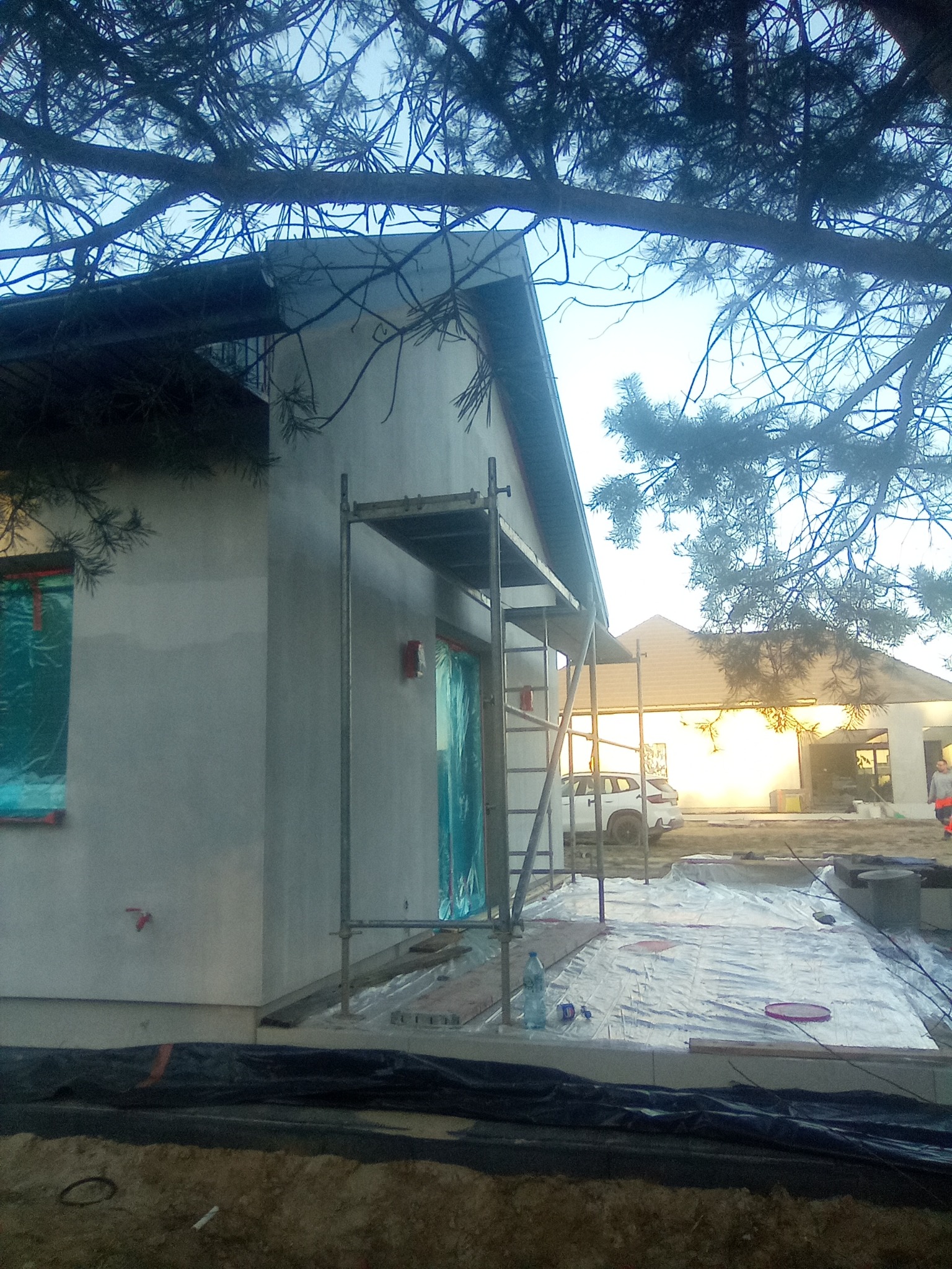 Dom w Piasecznie w trakcie tynkowania elewacji, widoczne rusztowania, okna zabezpieczone folią, w tle drugi budynek i samochód.