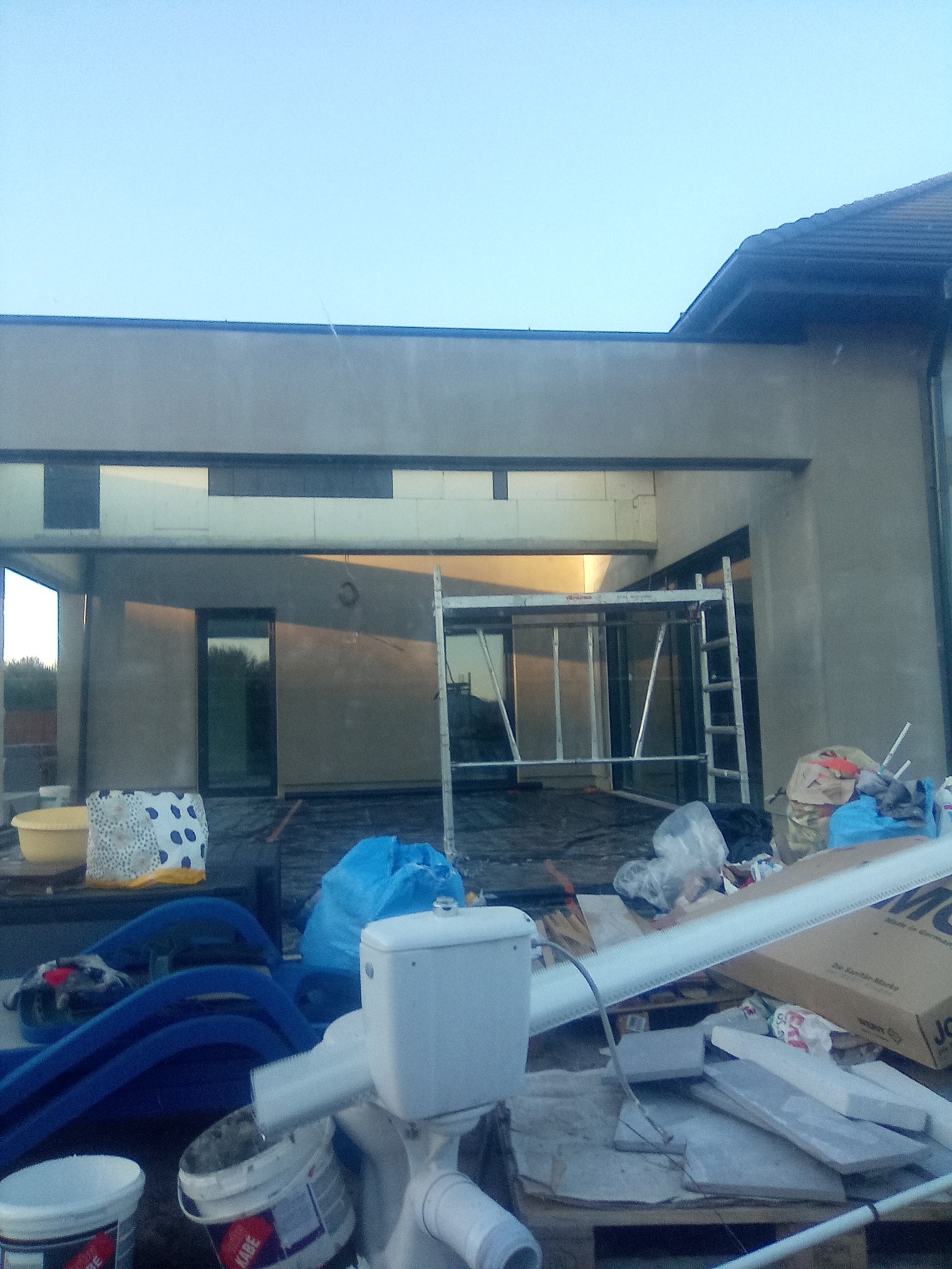 Surowy stan elewacji budynku w trakcie prac wykończeniowych, widoczne rusztowanie, ślady tynku i nieporządek na palecie z materiałami budowlanymi.