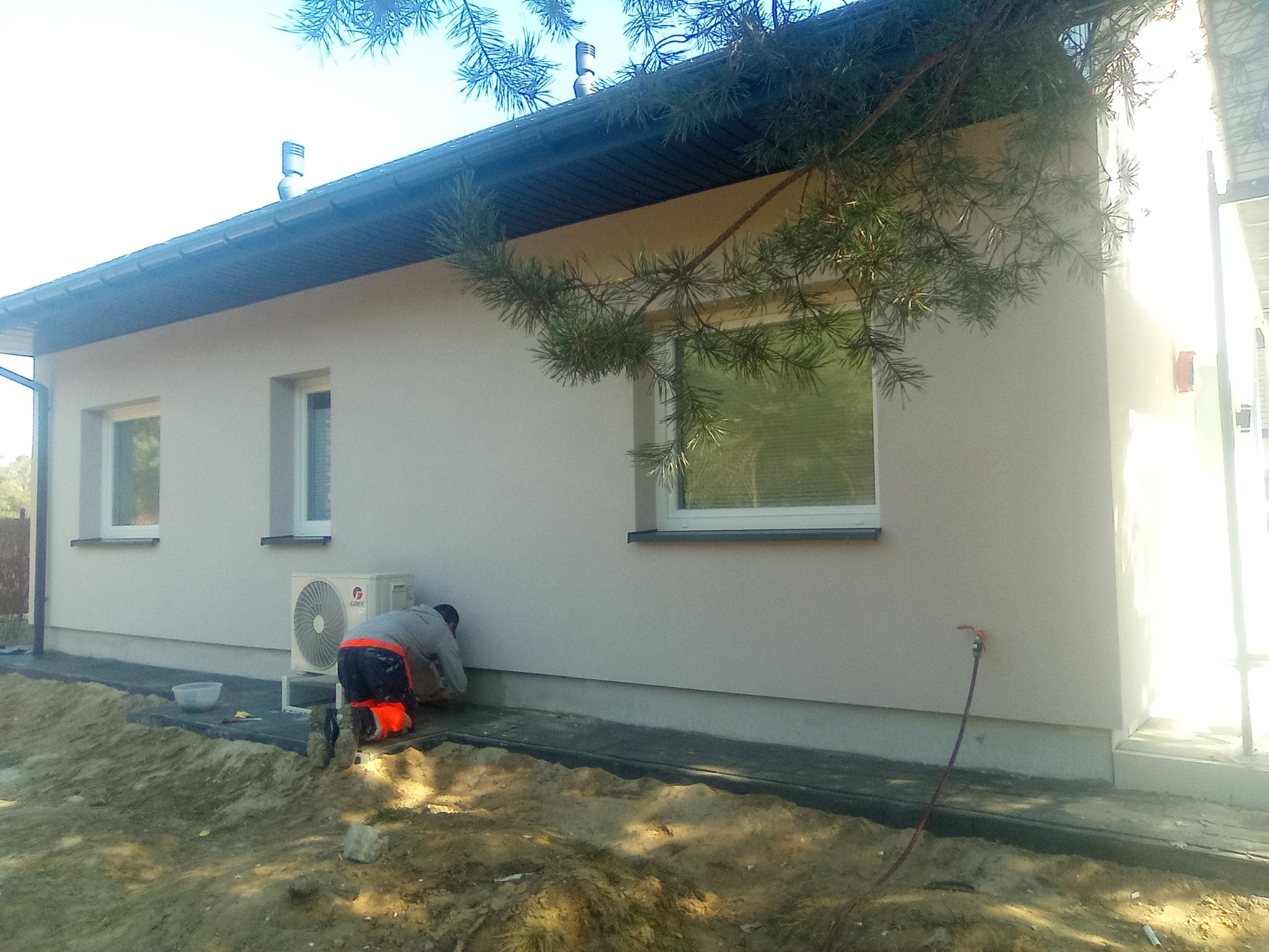 Pracownik w odzieży roboczej, klęczący przy ścianie domu z nową, jasną elewacją, wykonujący prace wykończeniowe przy cokole, widoczny klimatyzator na ścianie i elementy instalacji wodnej.
