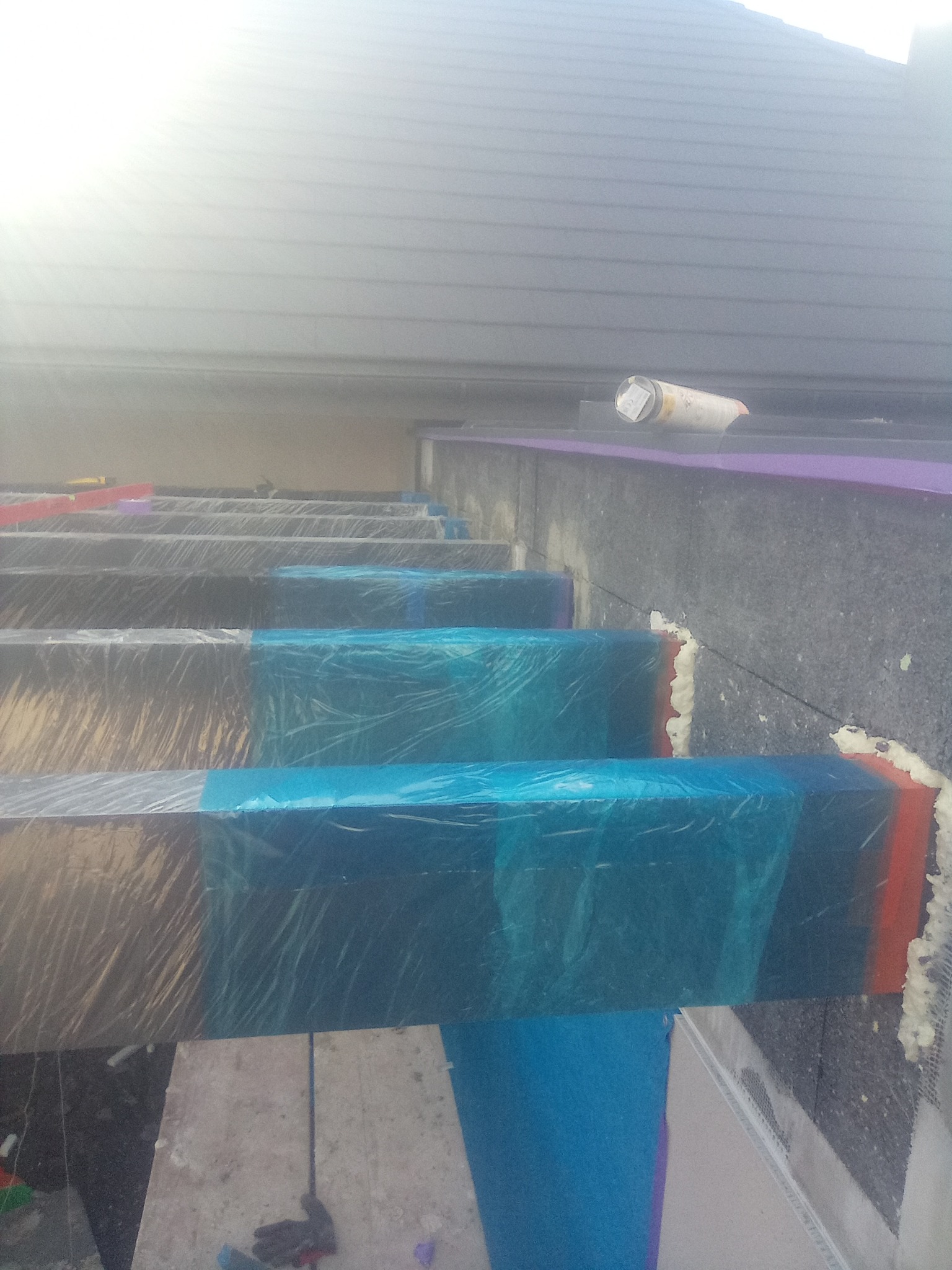 Montaż izolacji termicznej na elewacji budynku, widoczne płyty styropianowe zabezpieczone folią, pianka montażowa w szczelinach, częściowo widoczny dach budynku w tle.