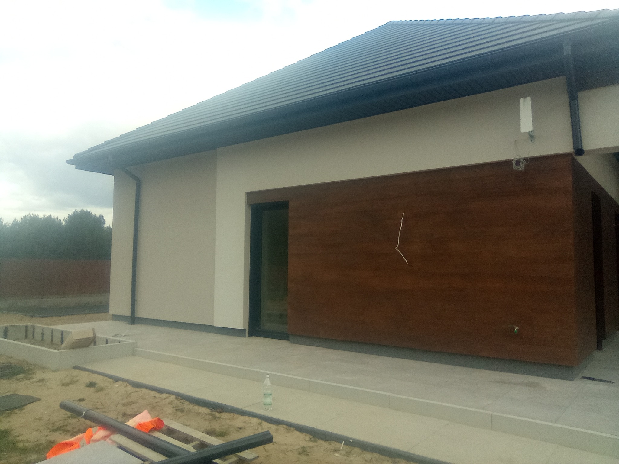 Nowoczesny dom jednorodzinny z jasną elewacją i panelami z drewna, widoczne elementy wykończeniowe, antena na ścianie, teren wokół domu w trakcie prac wykończeniowych.