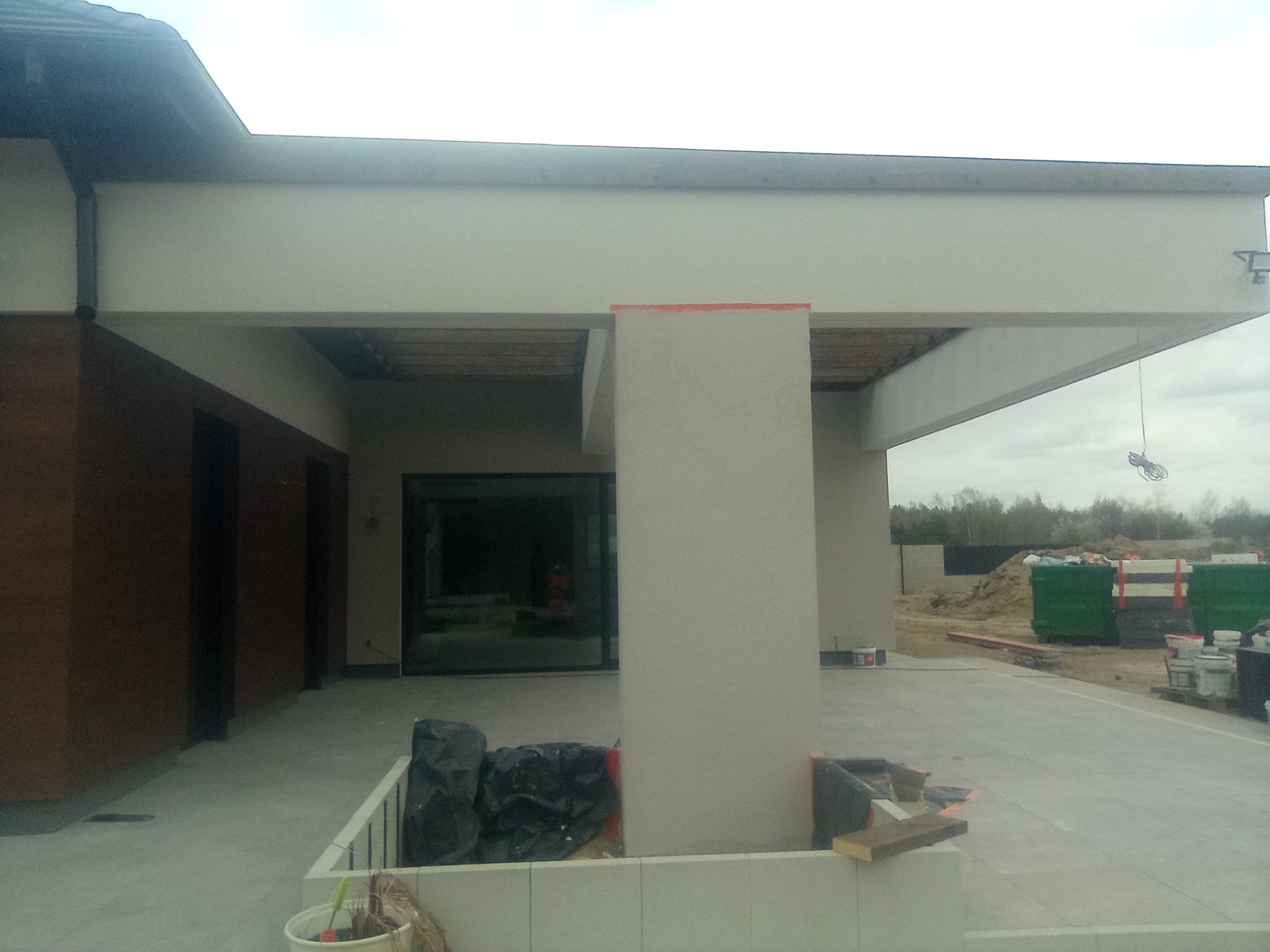 Nowoczesny dom w trakcie wykańczania elewacji, widoczne tynkowane filary, elementy drewniane pod dachem, teren budowy w tle z kontenerami.