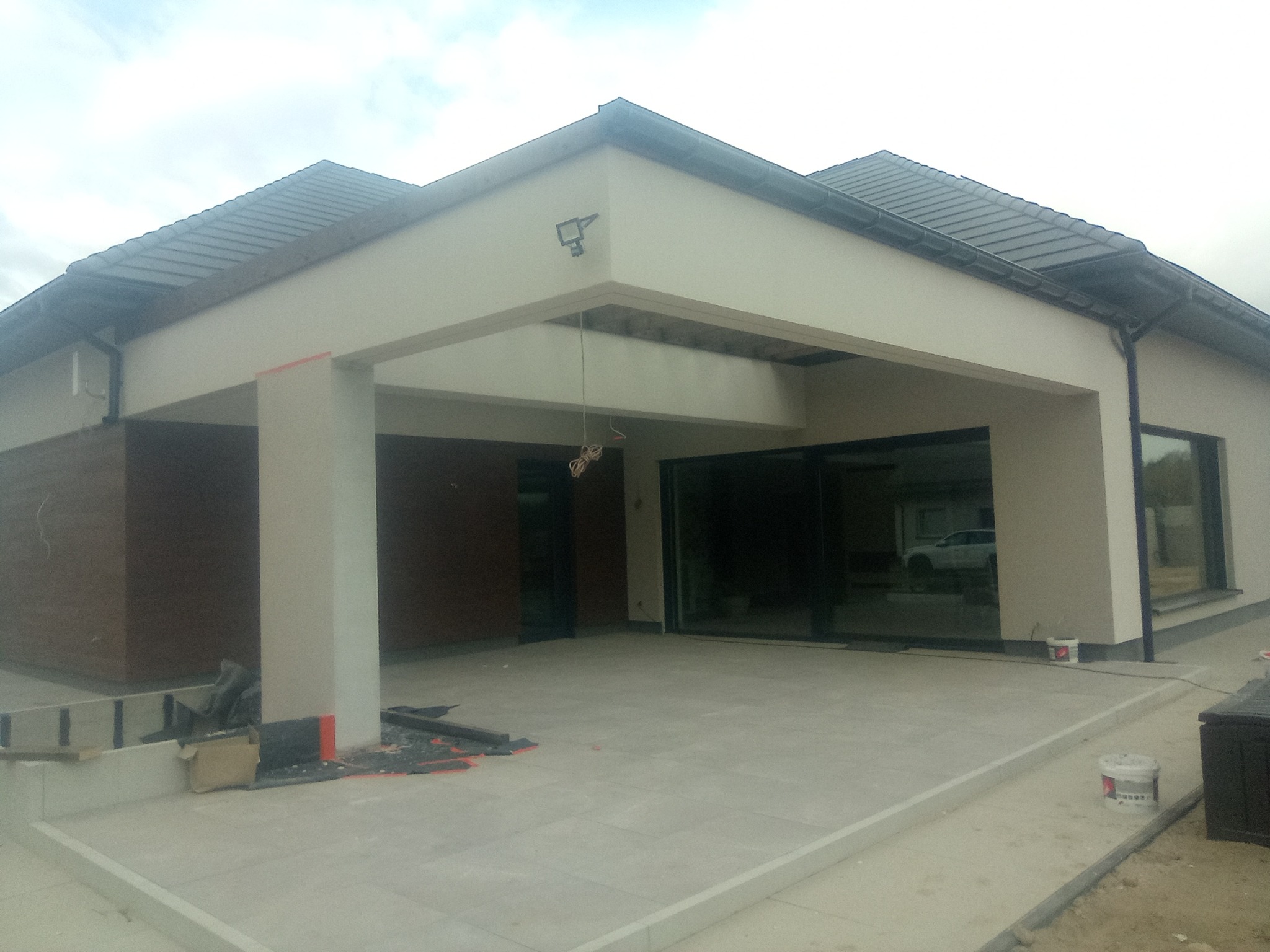 Nowoczesny dom jednorodzinny z beżową elewacją, widoczne elementy wykończeniowe, takie jak płytki na tarasie i fragmenty izolacji przy filarze. W tle zaparkowany samochód.