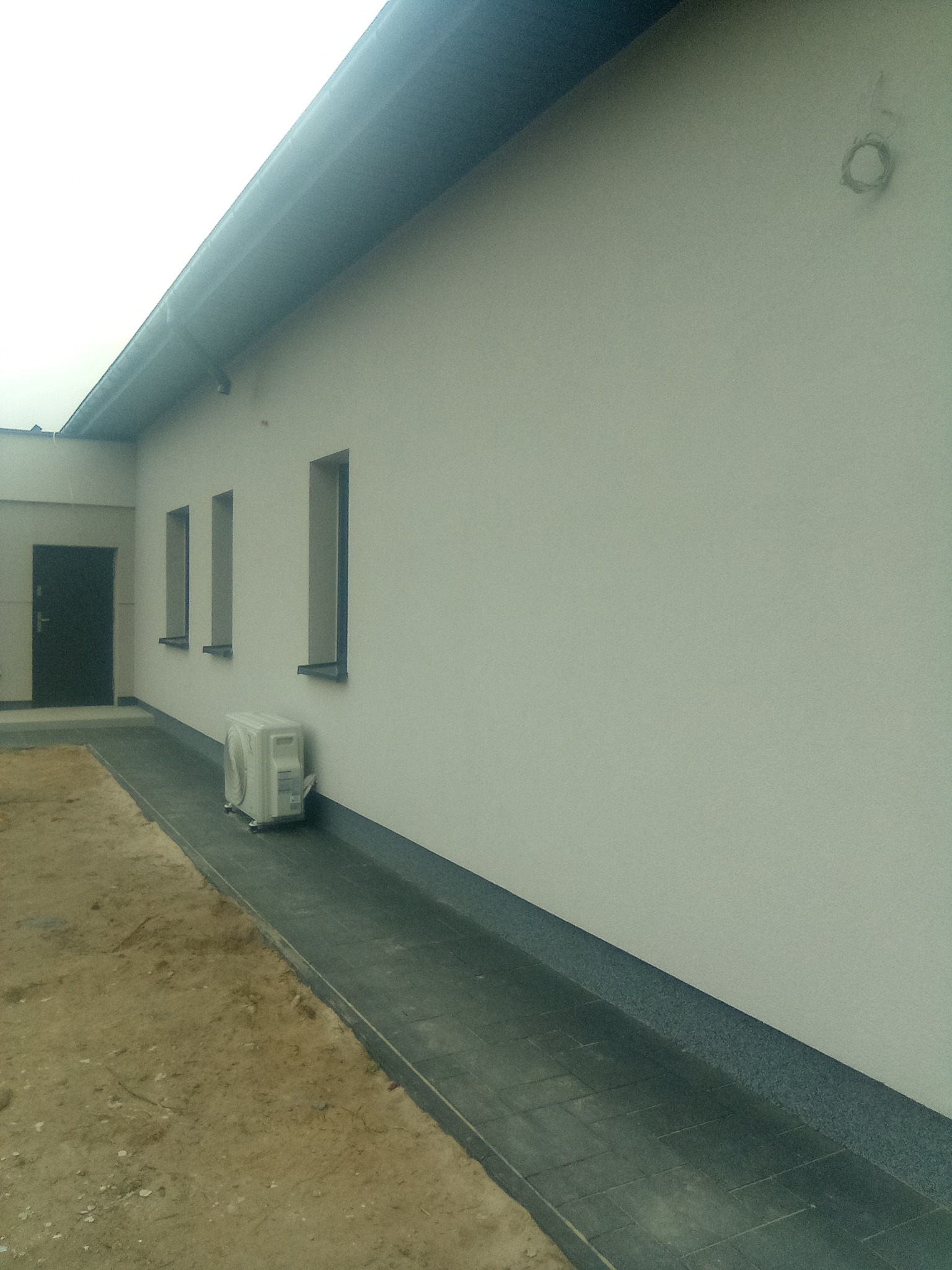 Boczny widok budynku z jasną, gładką elewacją, trzema wąskimi oknami i klimatyzatorem na zewnątrz, obok brukowanego chodnika.