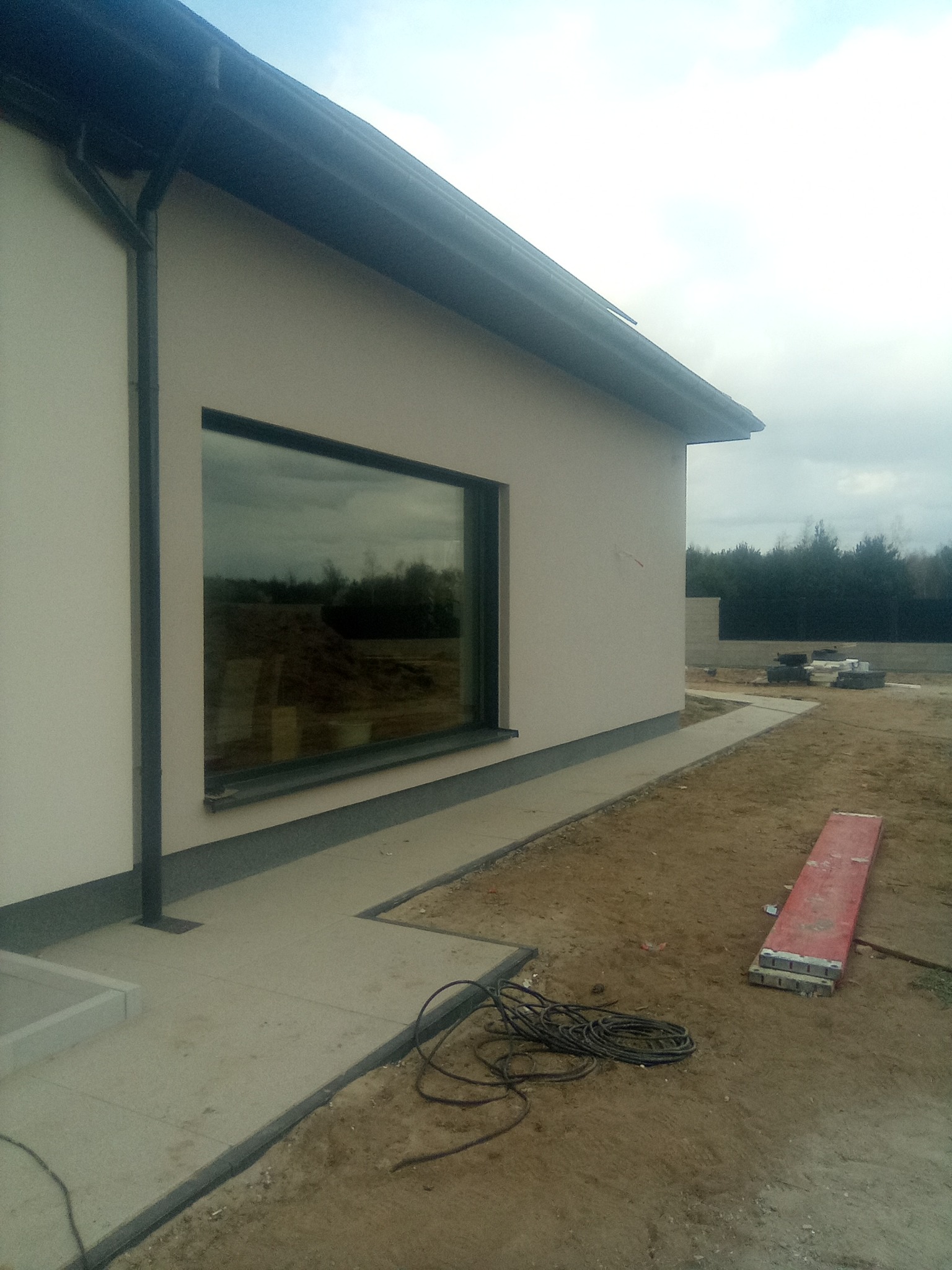 Fragment elewacji budynku z dużym oknem i betonową ścieżką wzdłuż ściany; na ziemi leżą zwinięte kable i czerwona deska.