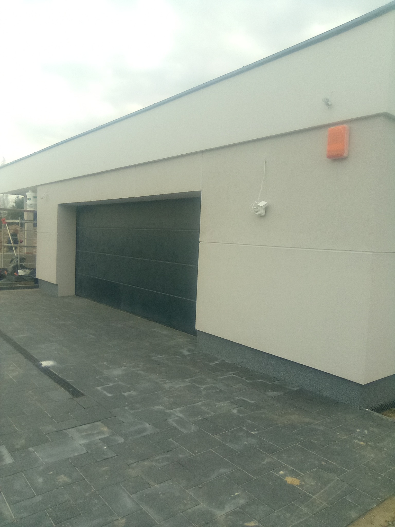 Elewacja budynku garażowego z szarą bramą segmentową, wykończona jasnym tynkiem strukturalnym oraz ciemnoszarą podmurówką i kostką brukową, widoczna pomarańczowa osłona na elewacji.