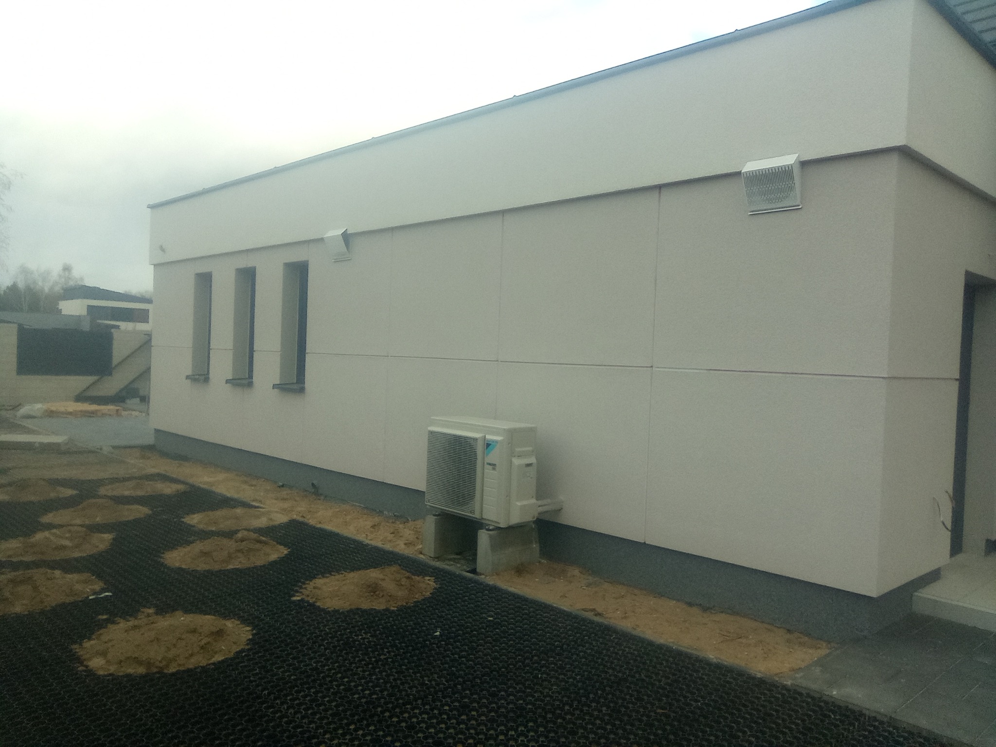 Jasna elewacja budynku z widocznym klimatyzatorem zewnętrznym na betonowych podstawkach, trawnik z kratki wypełniony ziemią, trzy wąskie okna, szara podmurówka i wentylator na ścianie.