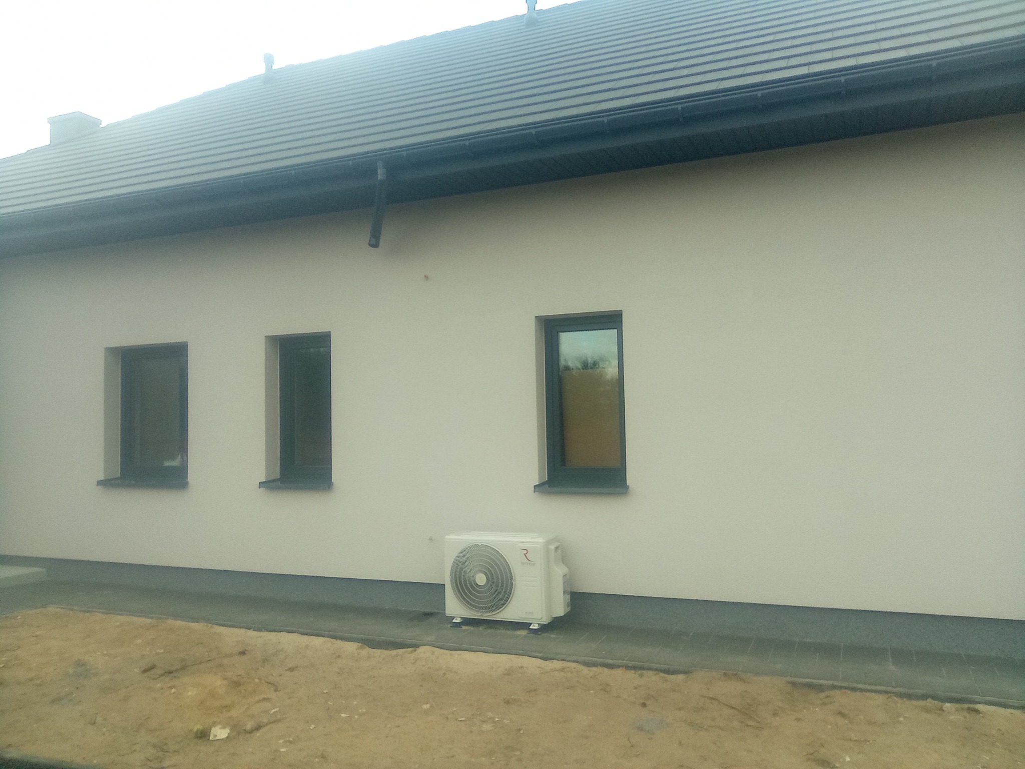 Otynkowana elewacja domu jednorodzinnego z trzema oknami i zamontowanym klimatyzatorem przy ścianie.