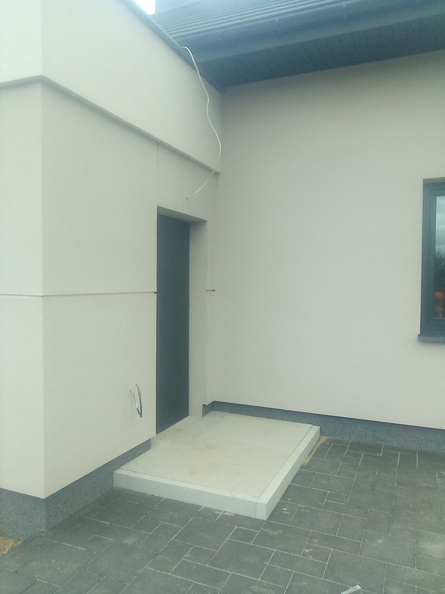 Jasna elewacja budynku z wejściem, widoczne elementy wykończeniowe oraz fragment okna, kostka brukowa przed wejściem.