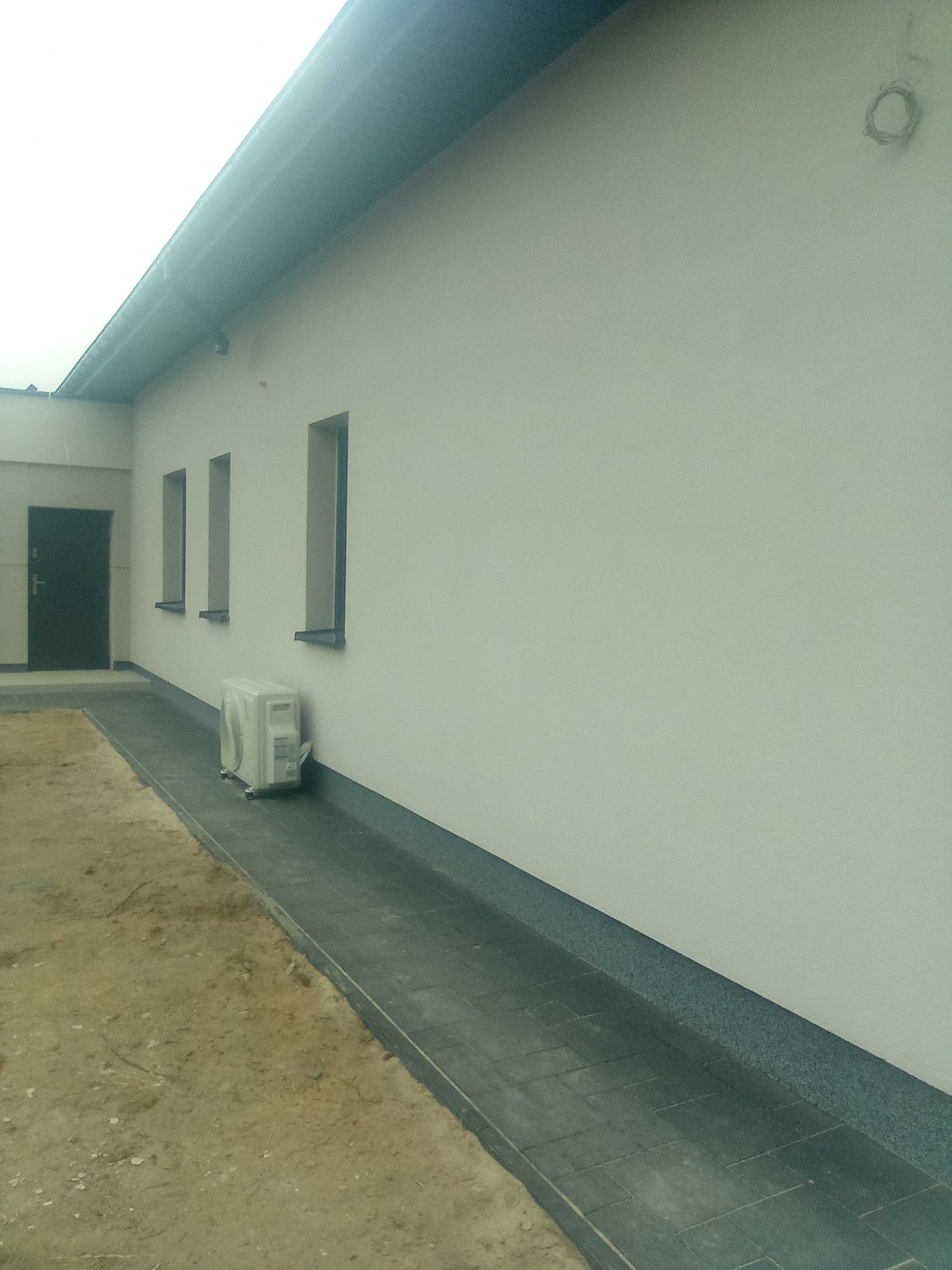 Jasna elewacja budynku z trzema wąskimi oknami, na tle czarna metalowa brama wejściowa, obok ściany stoi biały klimatyzator zewnętrzny, na dole szary chodnik i piasek.