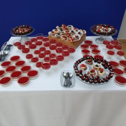 Liv Catering - Stół z deserami: mini tartaletki z owocami, pucharki z musem truskawkowym i napoje w kieliszkach. Biały obrus, niebieskie tło. Różnorodność słodkości.