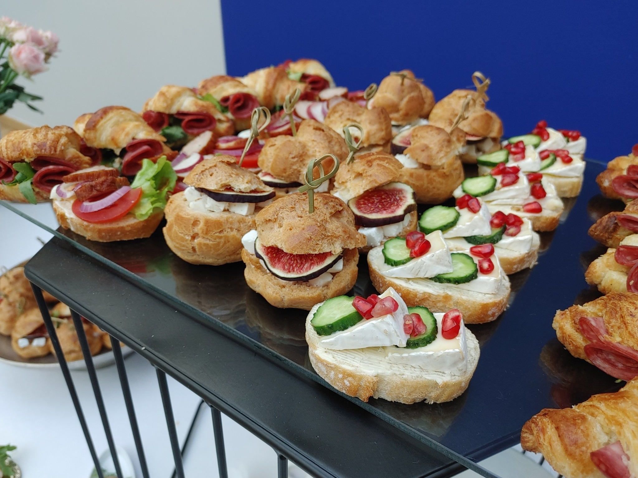 Elegancki catering: mini croissanty z szynką, kanapeczki z figą i pączki z kremem na czarnym, szklanym stojaku. Różowe kwiaty w tle dodają uroku.