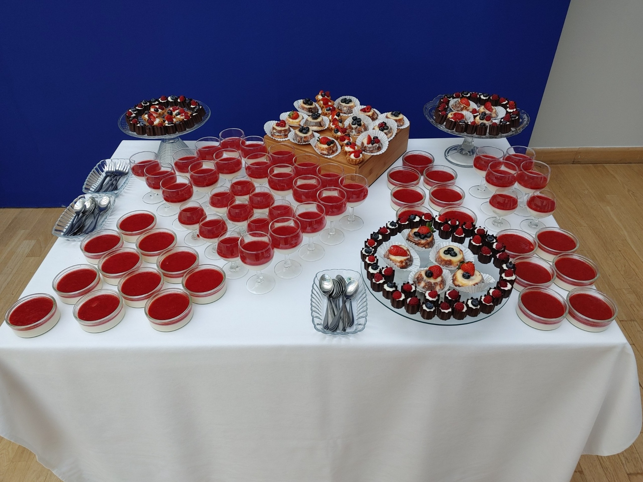 Stół z deserami: mini tartaletki z owocami, pucharki z musem truskawkowym i napoje w kieliszkach. Biały obrus, niebieskie tło. Różnorodność słodkości.