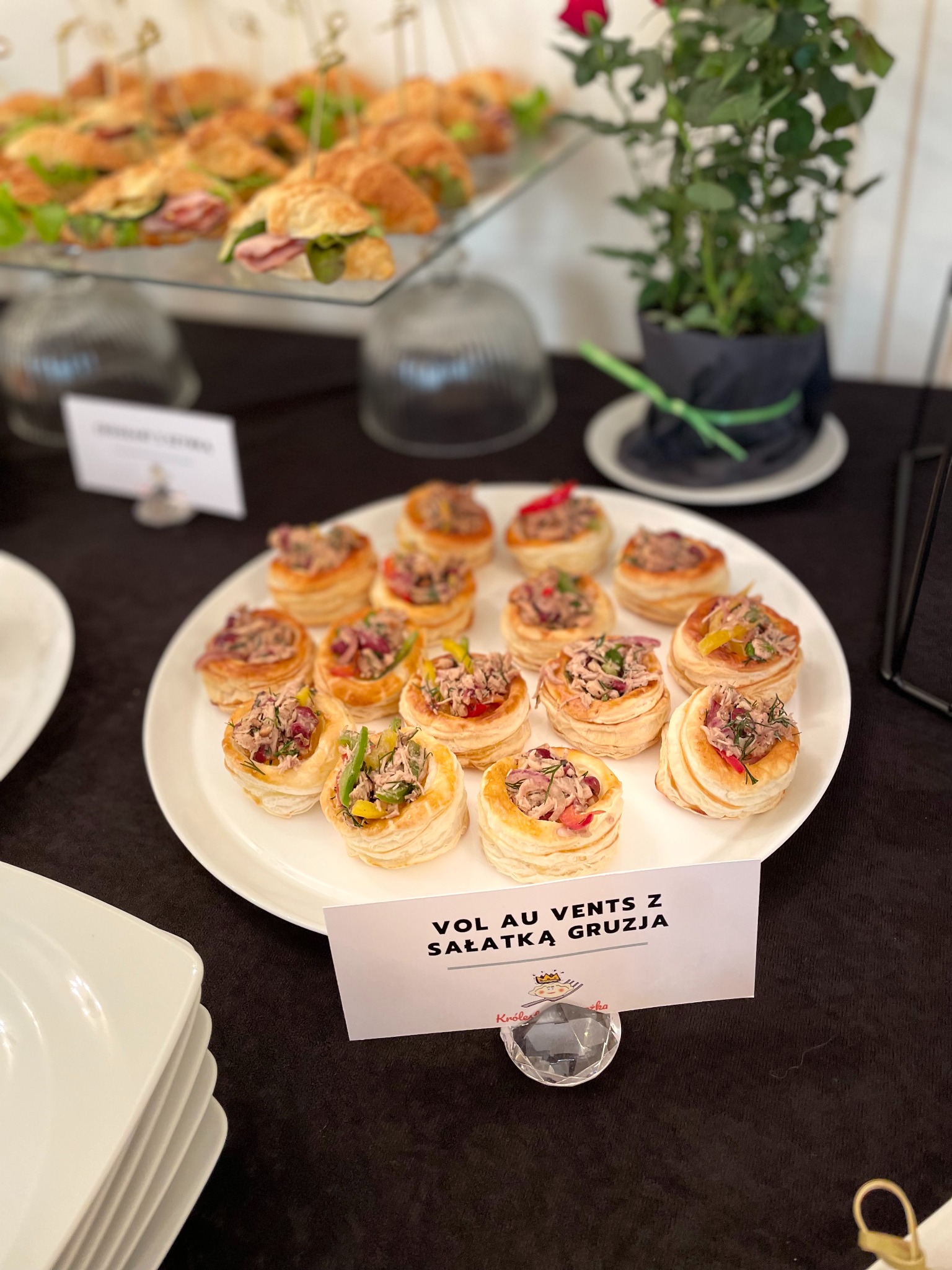 Elegancko podane vol au vents z sałatką gruzja na białym talerzu, obok kanapki z szynką i dekoracja z rośliny w czarnym kapeluszu. Catering.