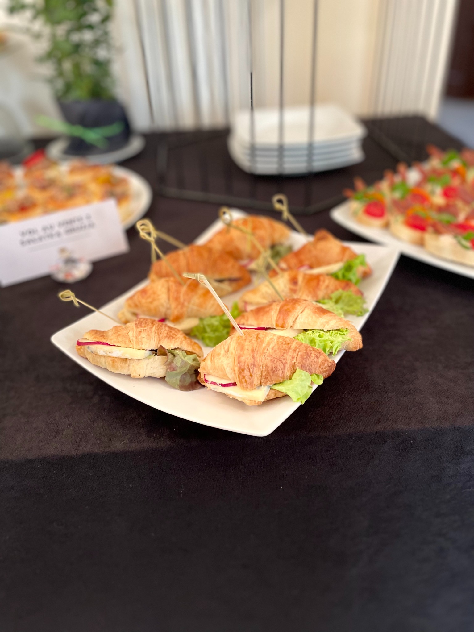 Kruche rogaliki z nadzieniem na białym talerzu, zabezpieczone ozdobnymi wykałaczkami. W tle inne przekąski i dekoracje na czarnym obrusie.