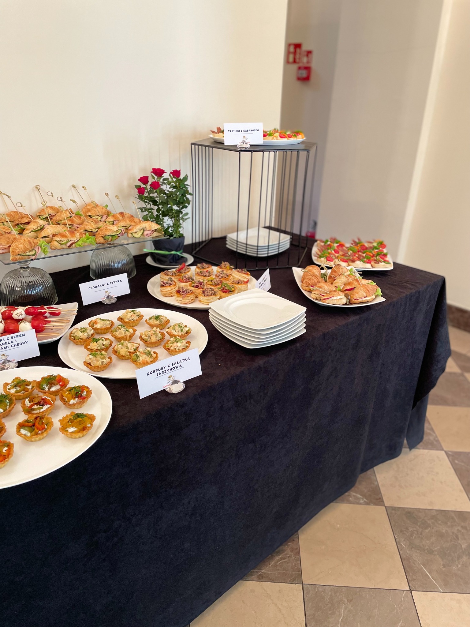 Stół cateringowy z różnorodnymi przekąskami: tartinki z kabanosem, korpusy z sałatką jarzynową, mini croissanty z szynką i szaszłyki z serem mozzarella i pomidorkami cherry.