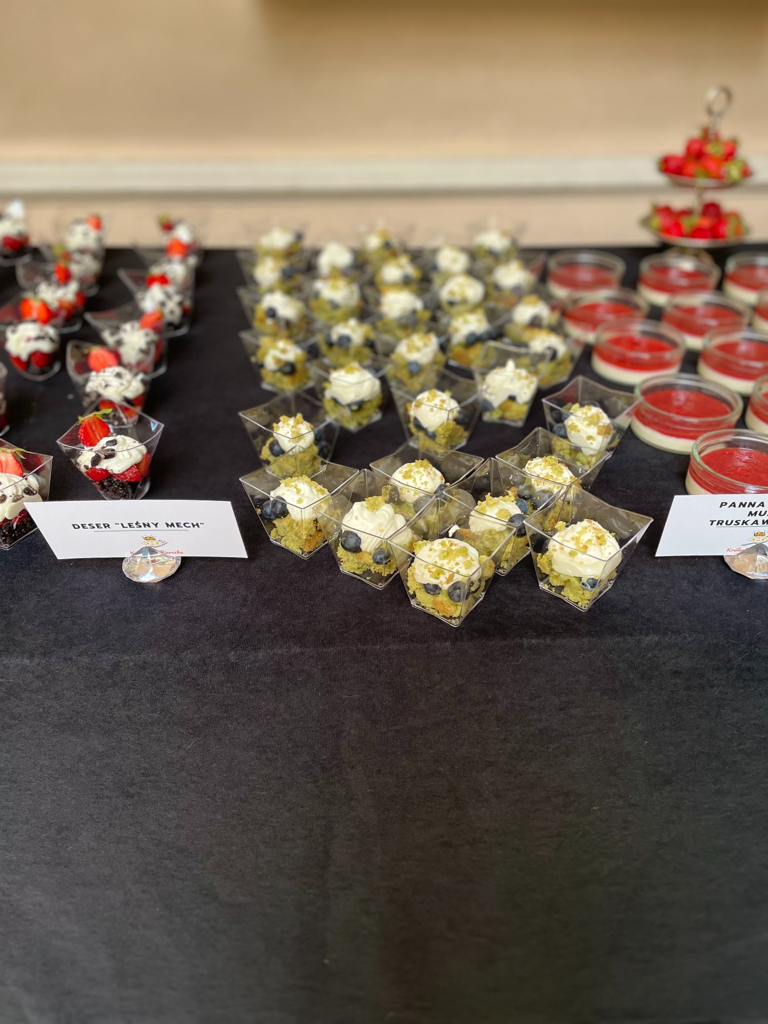 Desery w małych pojemnikach: leśny mech z borówkami, panna cotta truskawkowa. Elegancki catering deserowy na czarnym obrusie, z bliska.