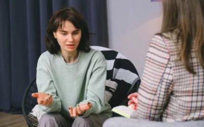 Młoda kobieta w jasnej bluzie siedzi na krześle z plecionki, gestykulując podczas rozmowy z osobą z notatnikiem, widoczną od tyłu w kraciastej marynarce.