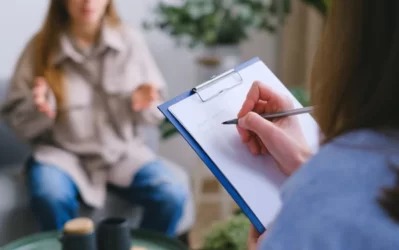 Osoba z długopisem wypełnia formularz na podkładce, w tle siedząca na kanapie kobieta w jasnej kurtce gestykuluje dłońmi.