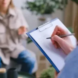 Psychoterapeuta robi notatki podczas sesji z pacjentką w jasnym, nowoczesnym gabinecie. Pacjentka siedzi na szarej sofie, ubrana w beżowy płaszcz i dżinsy.