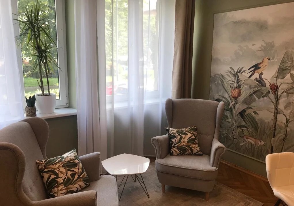 Jasny pokój z dwoma fotelami typu uszak z poduszkami w liście, małym białym stolikiem, rośliną doniczkową przy oknie i tapetą w motywy tropikalne z ptakiem.