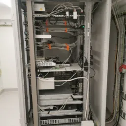 Otwarta szafa serwerowa z zainstalowanymi urządzeniami sieciowymi, widoczne kable uporządkowane pomarańczowymi opaskami, zasilacze UPS i listwy zasilające, okablowanie prowadzone w korytkach...