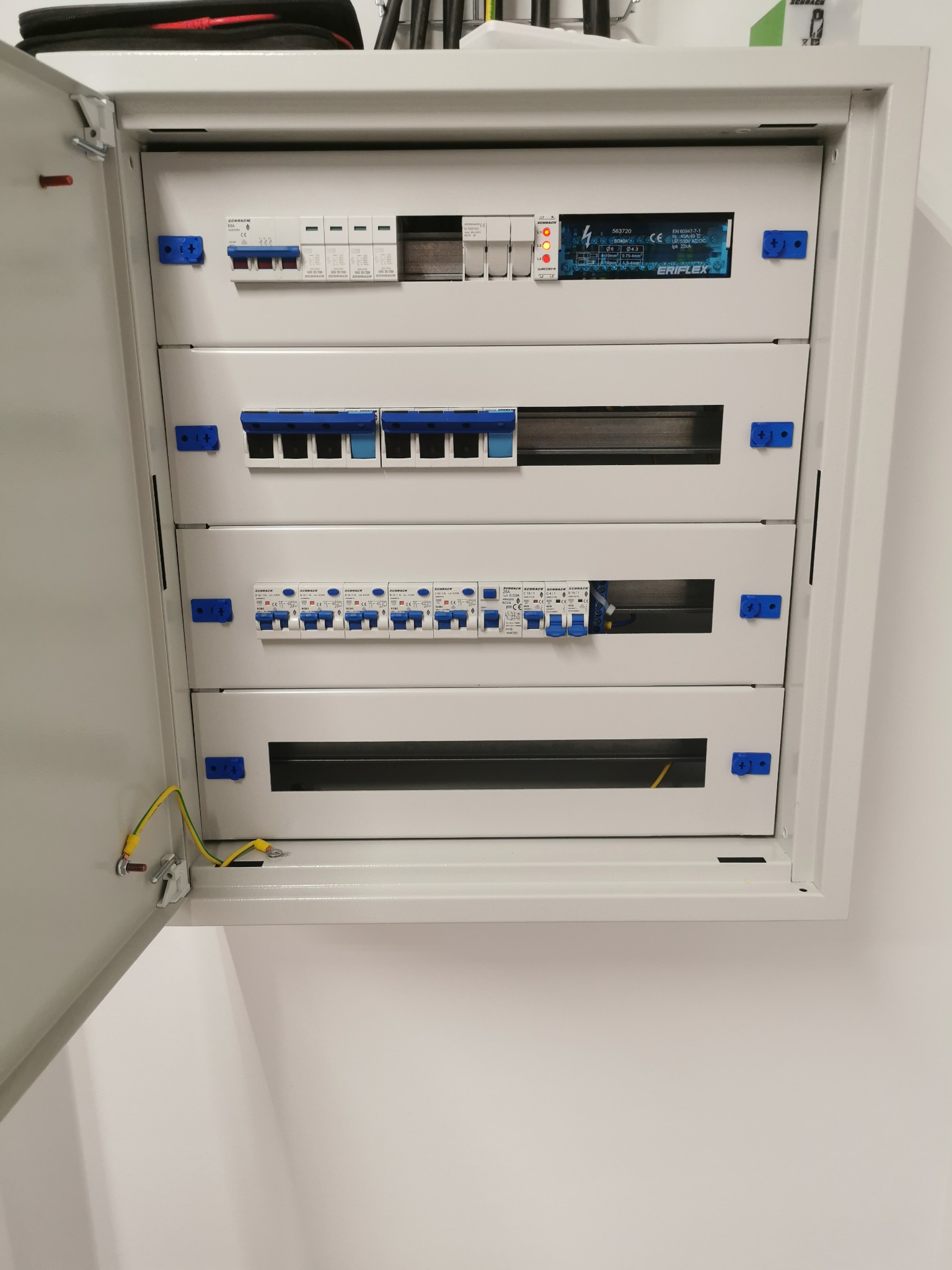 Otwarta skrzynka rozdzielcza z zainstalowanymi bezpiecznikami i zabezpieczeniami, widoczne przewody uziemiające przy drzwiach, elementy montażowe w kolorze niebieskim.